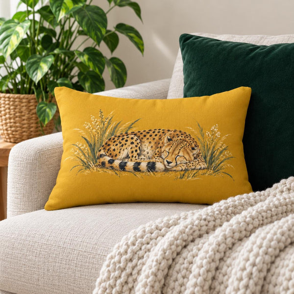 Mustard yellow lumbar pillow with sleeping cheetah and grass illustration styled on a cream sofa with dark green cushion, chunky knit blanket, and houseplant.
