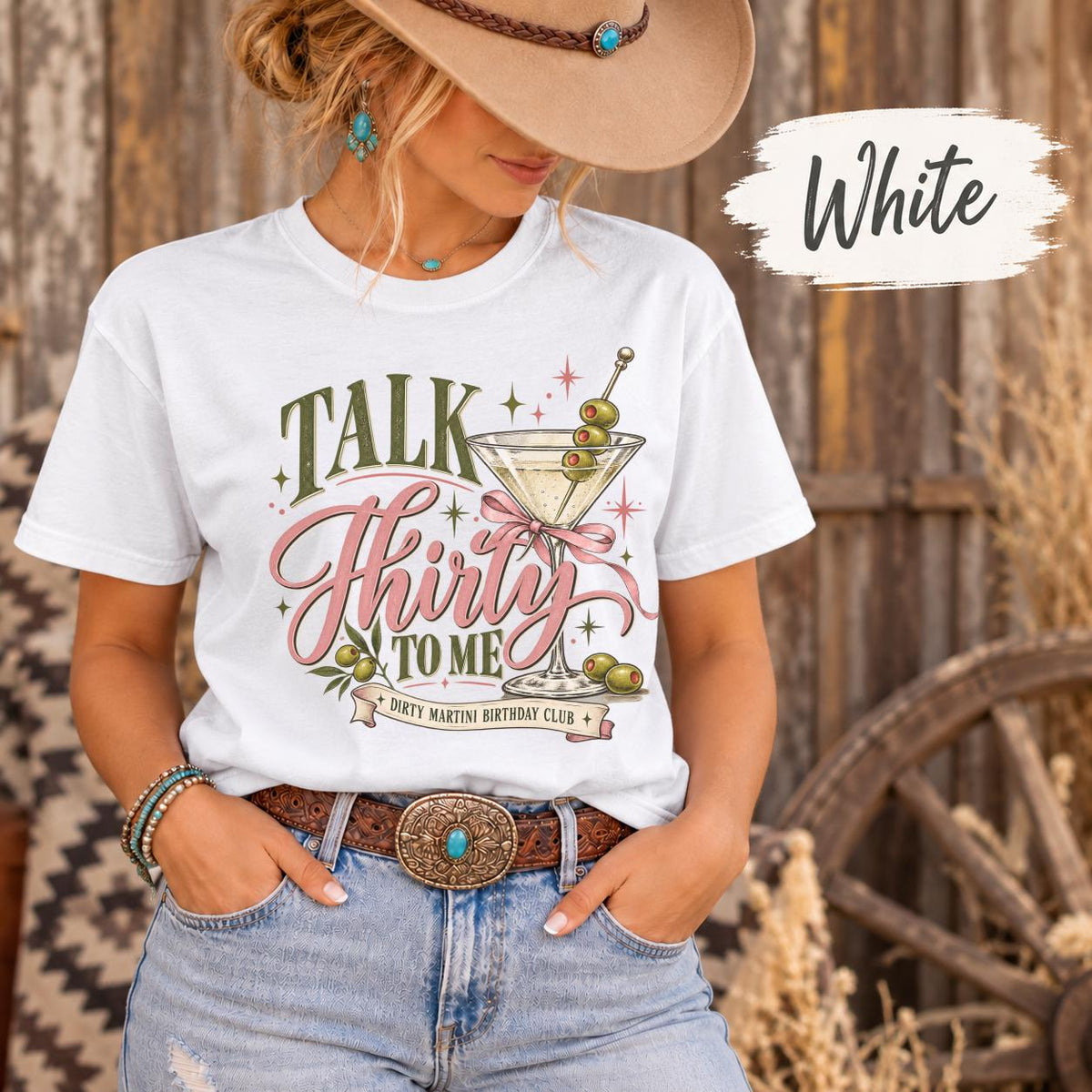 White T-shirt showcasing the dirty martini birthday design with pink and olive lettering, cocktail glass illustrations, and ribbon details.