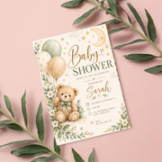 Editable baby shower invitation displayed on a blush-pink background with green botanical leaves. The charming watercolor artwork features a teddy bear, pastel balloons, golden celestial accents, and delicate flowers.