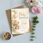 Gender-neutral baby shower invitation displayed on a pale blue background with pink flowers and leafy greenery. The watercolor design includes a sweet teddy bear, crescent moon, balloons, and golden stars.