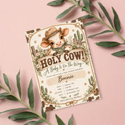 Holy Cow baby shower invitation displayed on a blush-pink background with green leafy branches. The country-inspired 5x7 design features a baby cow wearing a Western hat, white wildflowers, and a decorative rope frame.