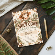 Holy Cow baby shower invitation displayed on a dark wooden tabletop with greenery and decorative accessories. The vintage country design features a cute calf, rope-style border, cream daisies, and rustic Western lettering.