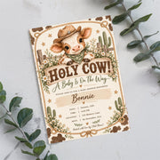 Printable Western baby shower invitation resting on a white background with eucalyptus branches. The farmhouse-inspired artwork features a sweet baby cow wearing a cowboy hat, botanical details, and rustic decorations.