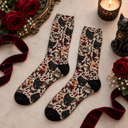 Pair of gothic cat crew socks displayed flat with deep red roses, burgundy velvet ribbon, a candle, and a black cat figurine. All-over vintage cat print features black, gray, and orange cats with floral accents.