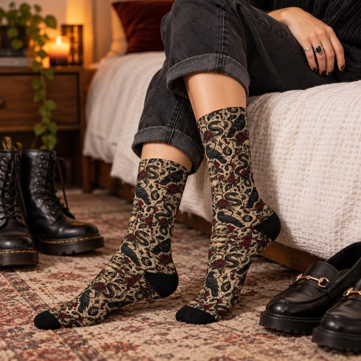 Gothic raven socks worn with cuffed black jeans in a cozy bedroom. Vintage-inspired all-over print features crows, coiled serpents, skulls, red roses, and moon details, styled with black boots nearby.