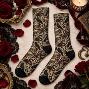 Pair of gothic raven crew socks displayed with burgundy roses, black lace, velvet ribbon, and antique decorations. Beige socks feature black ravens, winding snakes, skulls, celestial symbols, and contrasting black toes and heels.