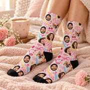 Custom face socks worn while relaxing on a soft pink blanket with a coffee mug and flowers nearby. Personalized design features repeating human and pet portraits with playful pink hearts, retro bows, and pastel accents.