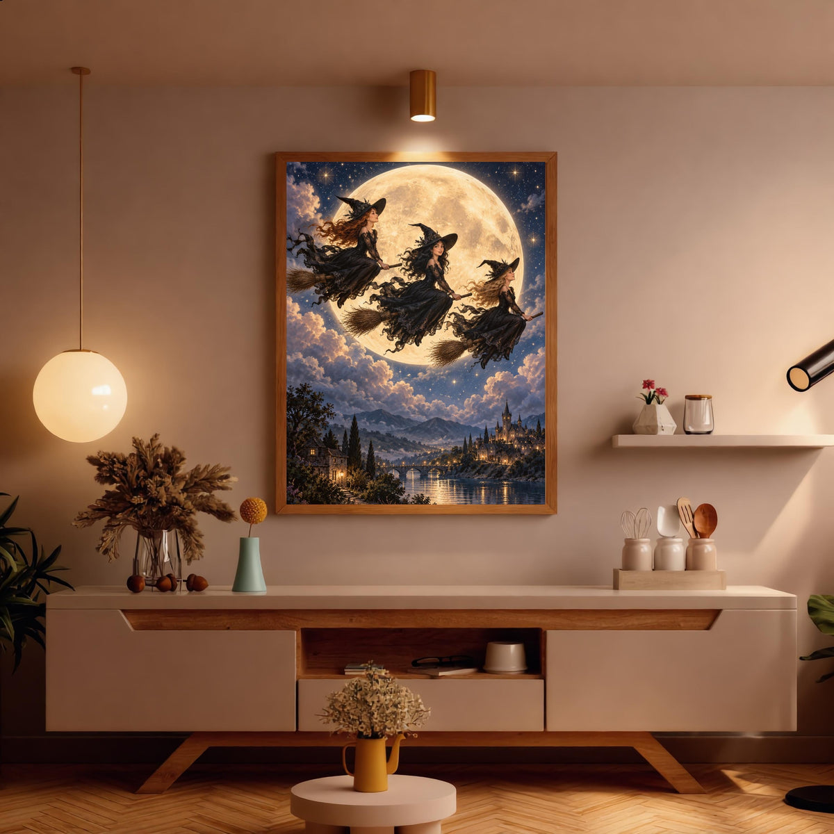 Witchy celestial art displayed above a wooden console in a warmly lit room. The illustration features flowing black dresses, three flying witches, a giant golden moon, and an enchanted riverside castle.