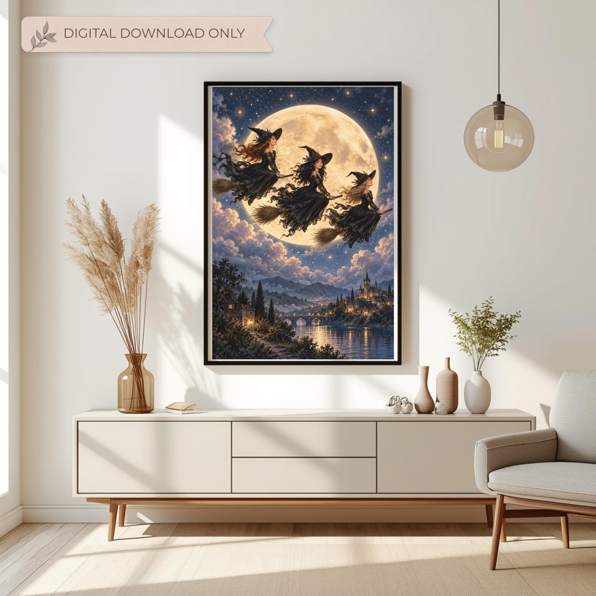 Framed witchy fantasy wall art displayed above a cream console table with dried botanical arrangements. The artwork features three witches flying on broomsticks across a golden full moon above an enchanted castle and moonlit river.