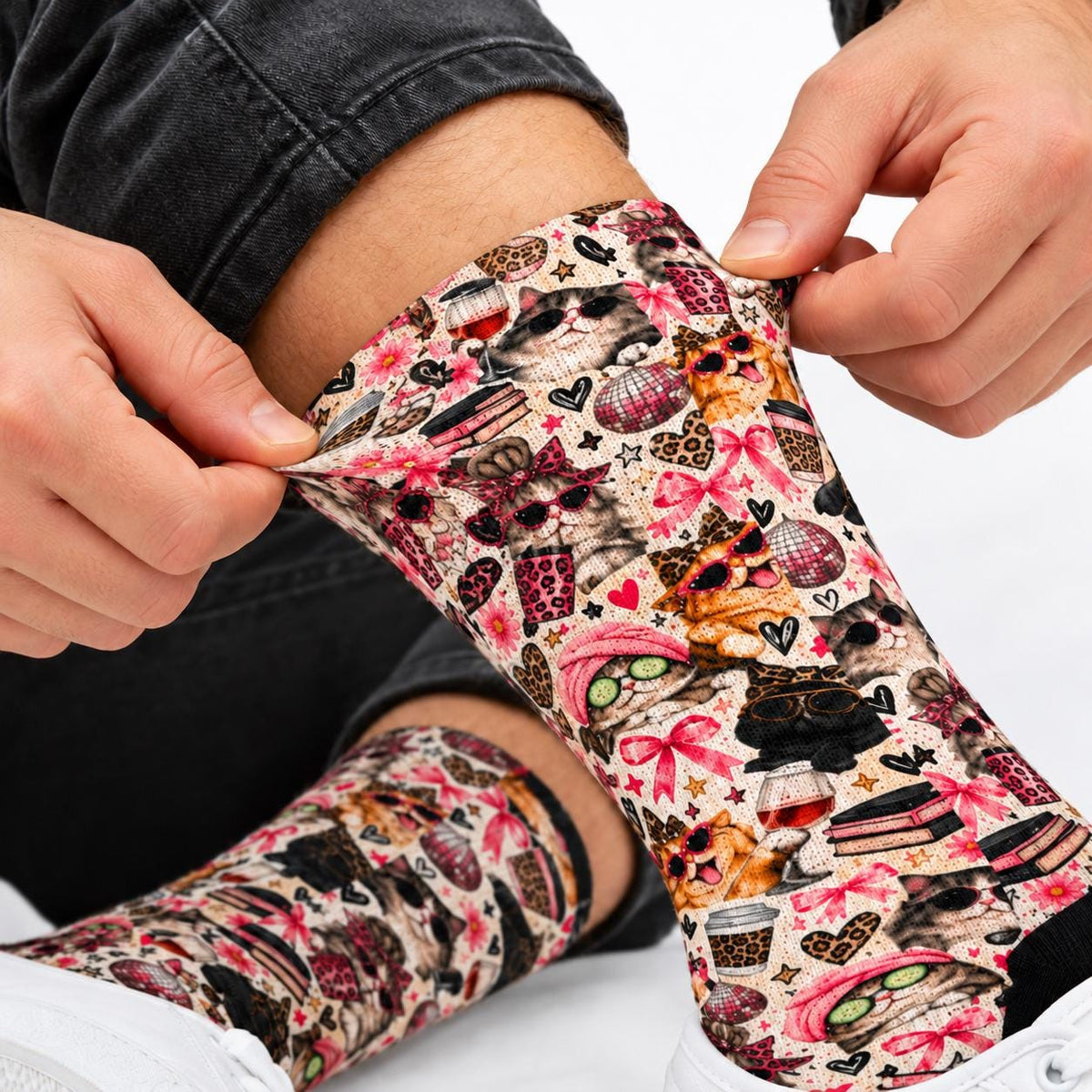 Close-up of retro kitty crew socks being gently stretched to display the printed fabric. Pattern features fashionable cats, pink bows, leopard print decorations, coffee cups, and playful hearts on a cream background.