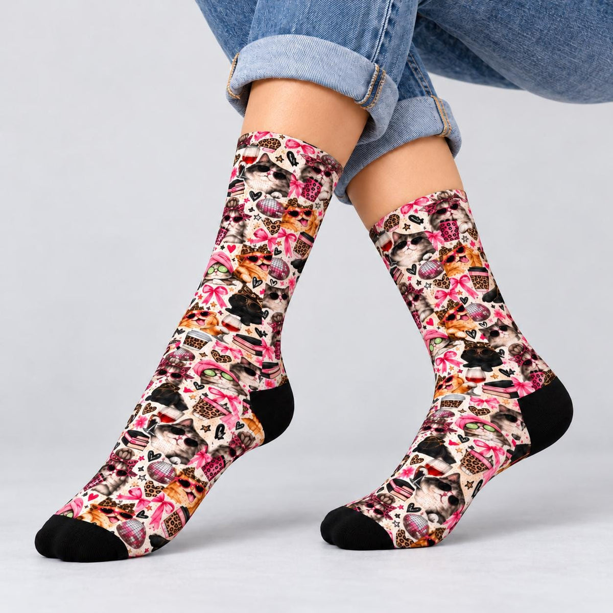 Pair of retro cat crew socks displayed on crossed legs against a neutral studio background. Whimsical print features stylish cats, pink bows, hearts, leopard accents, and contrasting black toes and heels.