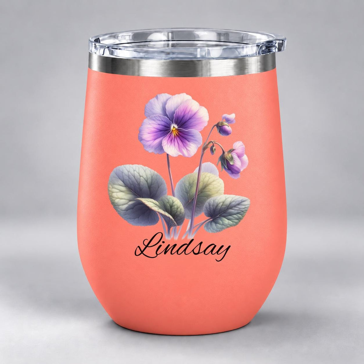 Coral stemless tumbler displaying the same violet floral artwork with personalized name on a powder-coated stainless steel cup.