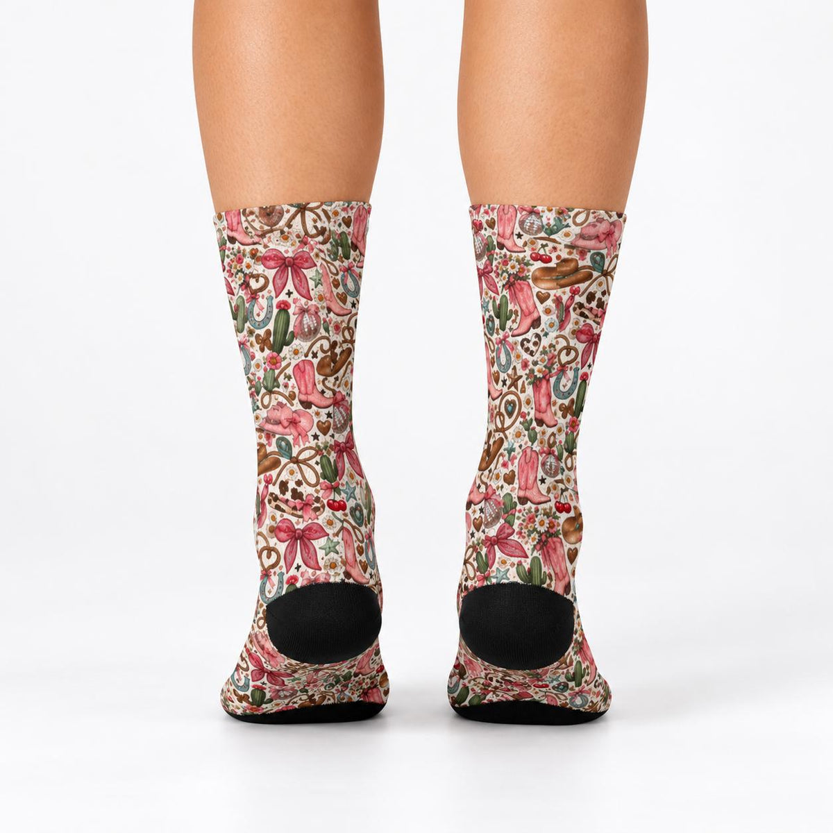 Rear view of women's cowgirl crew socks displaying the all-over vintage western pattern and black heels. Cream-colored socks feature pink boots, floral illustrations, horseshoes, bows, and green cactus accents.