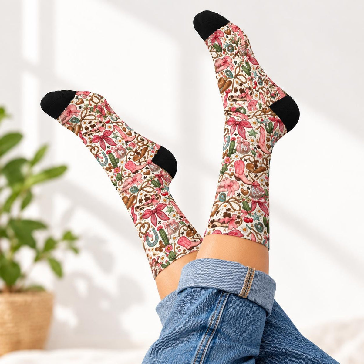 Retro western socks modeled with feet raised while wearing rolled blue jeans. Colorful design features pink cowboy boots, bows, cactus plants, cherries, hearts, and horseshoes with contrasting black toes and heels.
