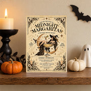 Vintage witch party invitation standing on a wooden shelf beside a black candlestick, orange pumpkin, and small white ghost. The Midnight Margaritas design features antique typography and magical celestial illustrations.