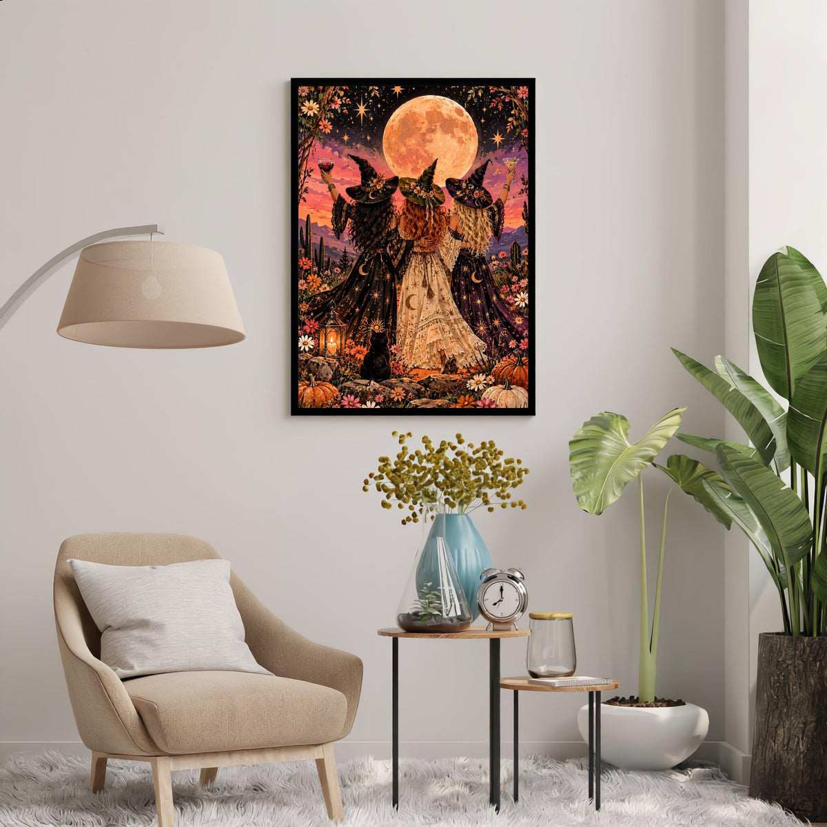 Retro celestial wall art displayed above a cream accent chair with a floor lamp and leafy plants. The whimsical poster features three witches celebrating friendship beneath a glowing moon with pumpkins and a black cat.