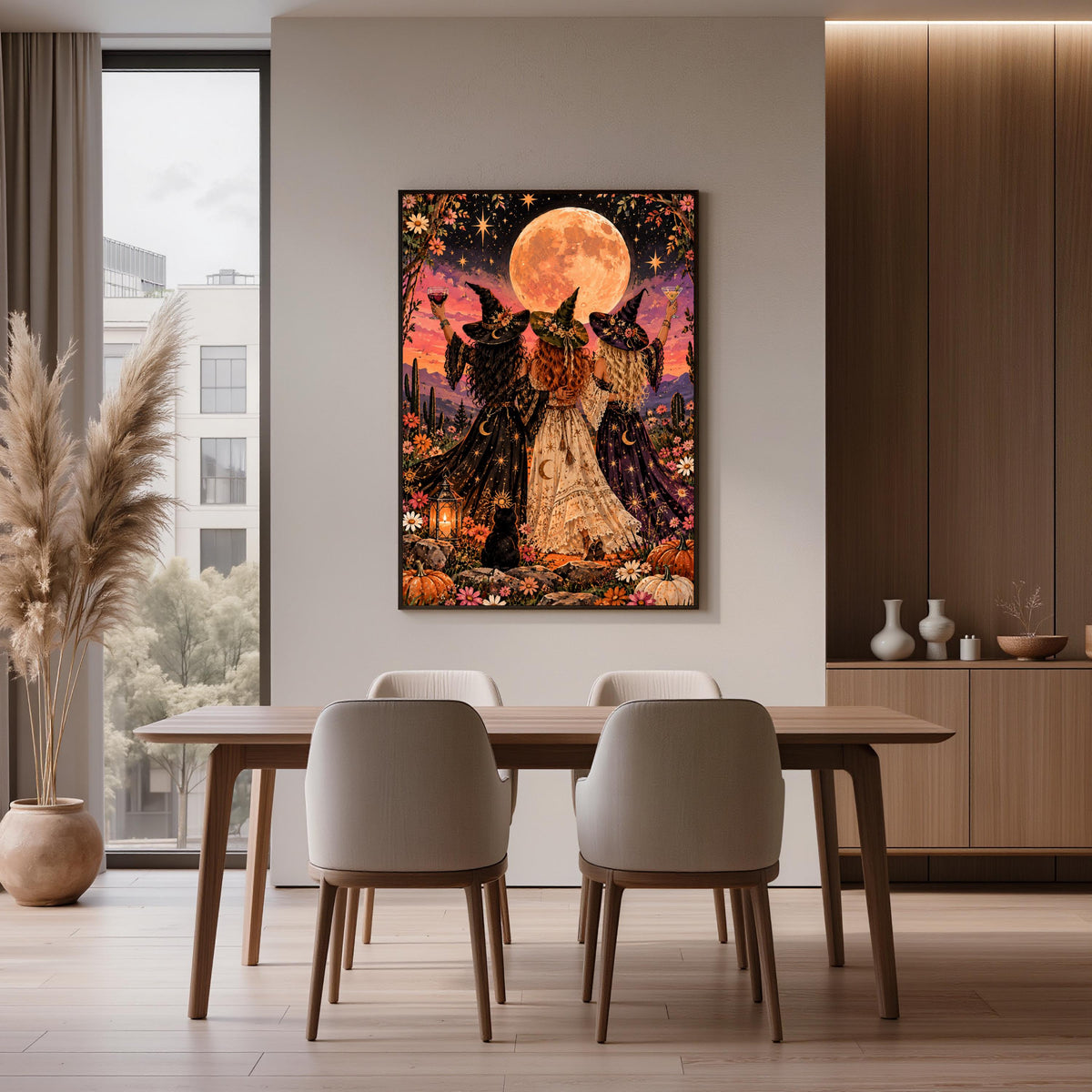Colourful Gothic boho wall art displayed above a wooden dining table in a contemporary interior. The fantasy illustration features three witches standing together beneath a golden moon, surrounded by flowers and celestial details.