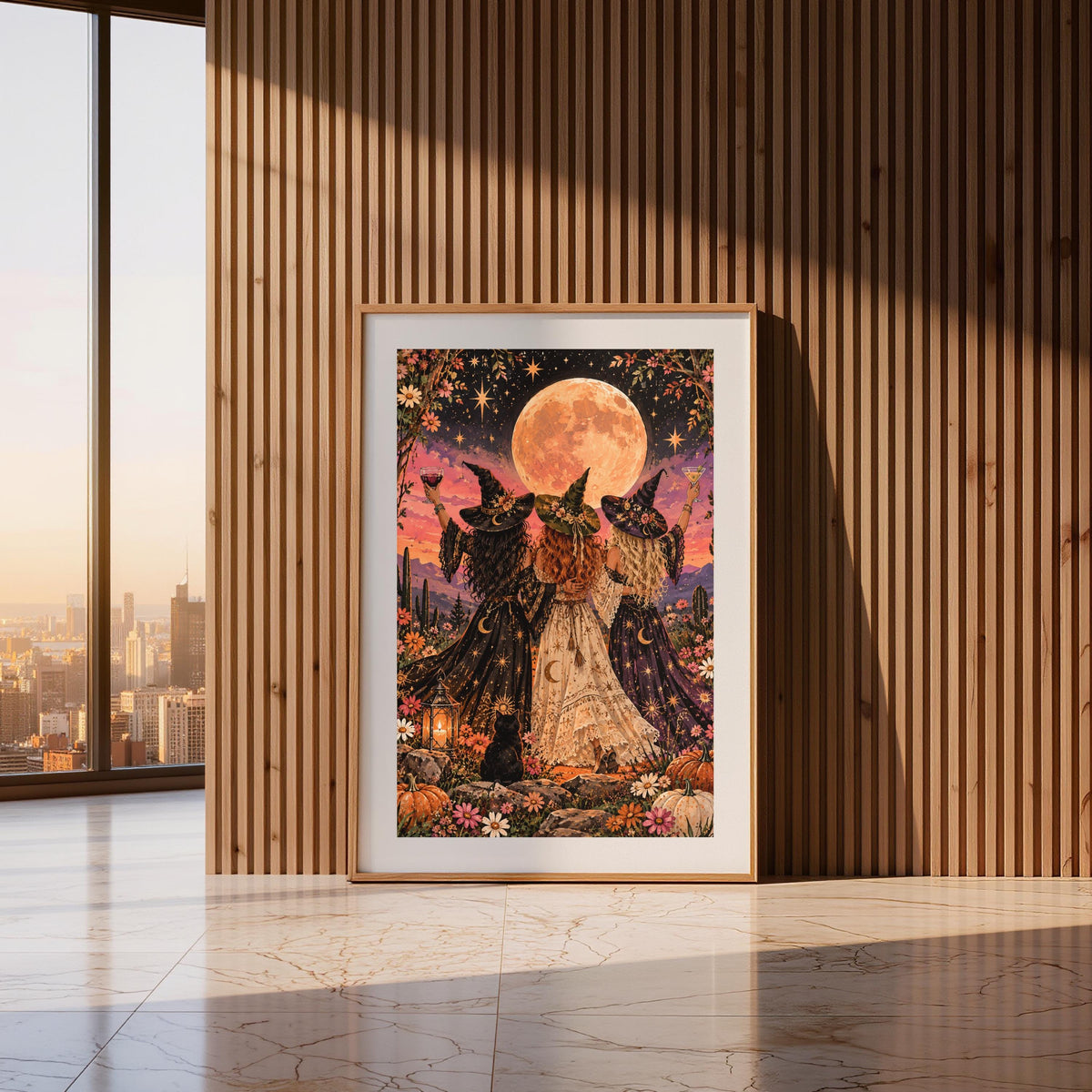 Framed whimsical Halloween poster displayed against a vertical wood-slat wall in a warm modern hallway. The colourful illustration features three witches in pointed hats standing beneath a giant peach moon.