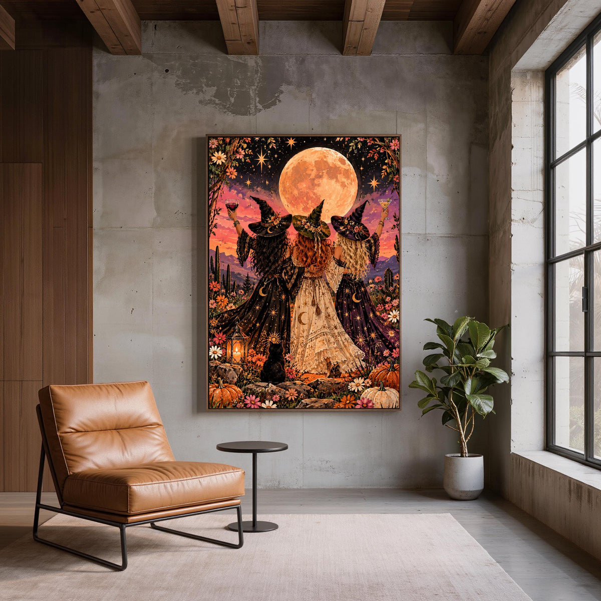 Moonlight, Magic & Mischief wall art displayed on a textured grey wall beside a tan leather chair. The mystical poster features three witches celebrating beneath a full moon with desert flowers and a black cat.