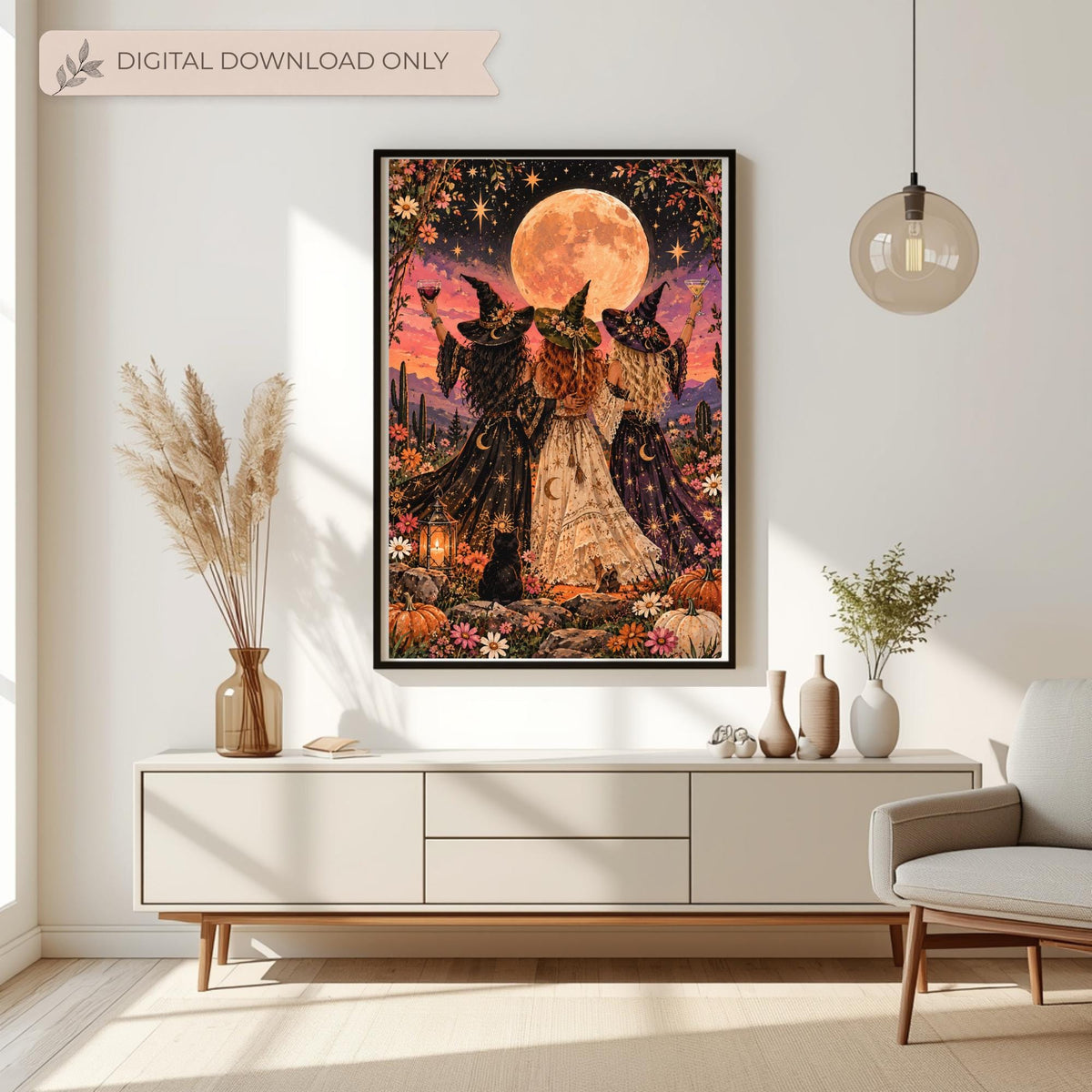 Framed retro witchy wall art displayed above a cream console table with dried botanical arrangements. The colourful fantasy print features three witches raising their glasses beneath a glowing peach moon, surrounded by wildflowers, pumpkins, and a black cat.