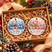 Pair of blue and pink Baby’s First Christmas ornaments nestled in kraft gift packaging with festive greenery and pinecones.