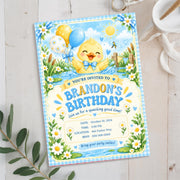 Cute duckling birthday invitation displayed on a white surface surrounded by delicate flowers and greenery, featuring pastel blue and yellow artwork for a little boy's birthday celebration.