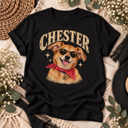 Black T-shirt flat lay showing the personalized Chester dog illustration with cream vintage lettering, sunglasses, red bandana and distressed graphic details in a coordinated boho setting.