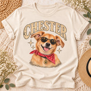 Ivory T-shirt mockup featuring Chester custom dog portrait artwork with a golden dog wearing sunglasses and a red bandana, displayed on a neutral background with flowers and woven accessories.