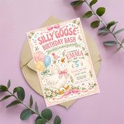 Two-sided Silly Goose birthday invitation arranged on a pastel pink surface with eucalyptus leaves, featuring whimsical goose artwork, floral details and pink gingham decorations.