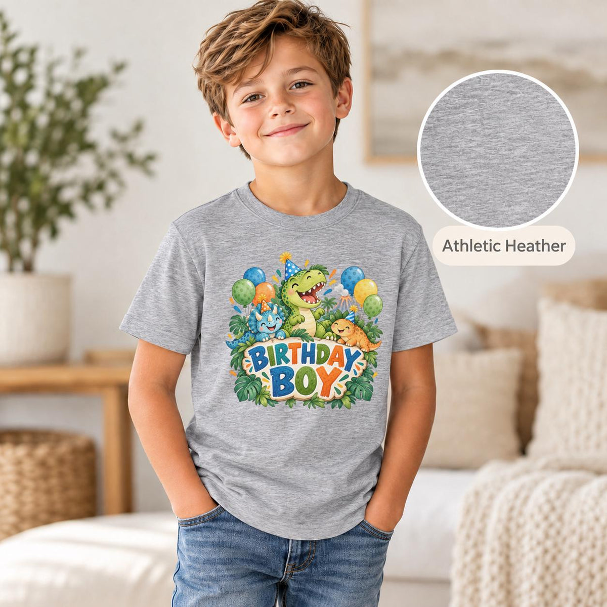 Boy wearing a sport athletic heather birthday graphic tee featuring three happy dinosaurs in party hats, blue and orange balloons, green jungle foliage and colorful lettering.