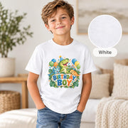Smiling young boy wearing a white dinosaur birthday T-shirt with three cheerful cartoon dinosaurs, colorful balloons, party hats and tropical leaves.