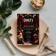 Editable Dirty Thirty birthday invitation resting on soft beige fabric beside botanical leaves and decorative accessories. Glamorous martini-themed 30th birthday party design with red and gold details.