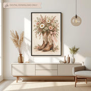Framed cowgirl wall art featuring vintage embroidered cowboy boots filled with pink and white wildflowers and a gingham bow. Displayed above a cream console table with dried botanical decorations in a bright neutral living room.