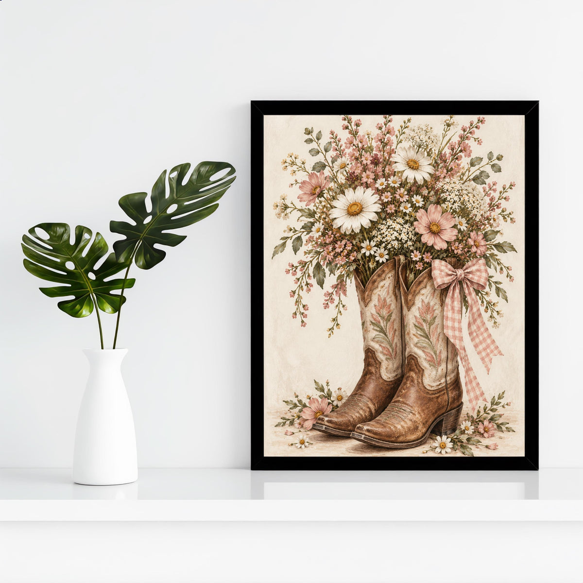 Vintage cowboy boots printable displayed in a black frame against a clean white wall beside a small green plant. The artwork combines brown leather boots, delicate pink flowers, and a soft ivory background.