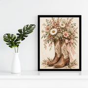 Vintage cowboy boots printable displayed in a black frame against a clean white wall beside a small green plant. The artwork combines brown leather boots, delicate pink flowers, and a soft ivory background.