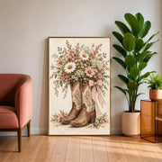 Pink cowgirl wall art resting on a wooden cabinet beside a large green houseplant and upholstered accent chair. The floral boot illustration features blush daisies, cream blossoms, and a gingham ribbon.