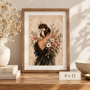 8 x 12 inch romantic floral portrait displayed in a wooden frame on a light console table with neutral home accessories. The museum-grade matte poster features a woman viewed from behind wearing a black dress and ivory bow, surrounded by pink and white botanical flowers.