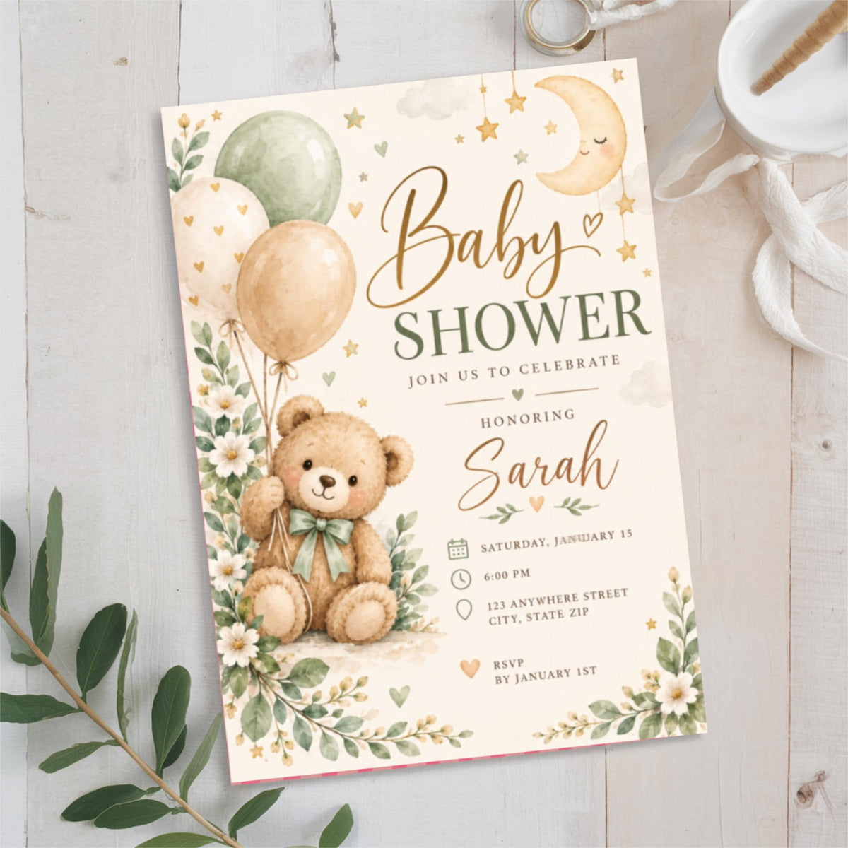 Watercolor baby shower invitation displayed on soft white fabric with eucalyptus leaves. The gender-neutral design features a cuddly teddy bear holding balloons, sage green accents, scattered stars, and floral illustrations.