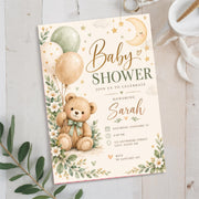 Watercolor baby shower invitation displayed on soft white fabric with eucalyptus leaves. The gender-neutral design features a cuddly teddy bear holding balloons, sage green accents, scattered stars, and floral illustrations.