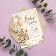 Editable teddy bear baby shower invitation resting on a blush-pink background surrounded by leafy greenery. The whimsical design features pastel balloons, golden stars, a sleepy crescent moon, and delicate white flowers.