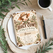 Western baby shower invitation displayed on a round white tray surrounded by botanical greenery and neutral decor. The rustic design features an adorable baby calf, cowboy hat, vintage lettering, and floral illustrations.