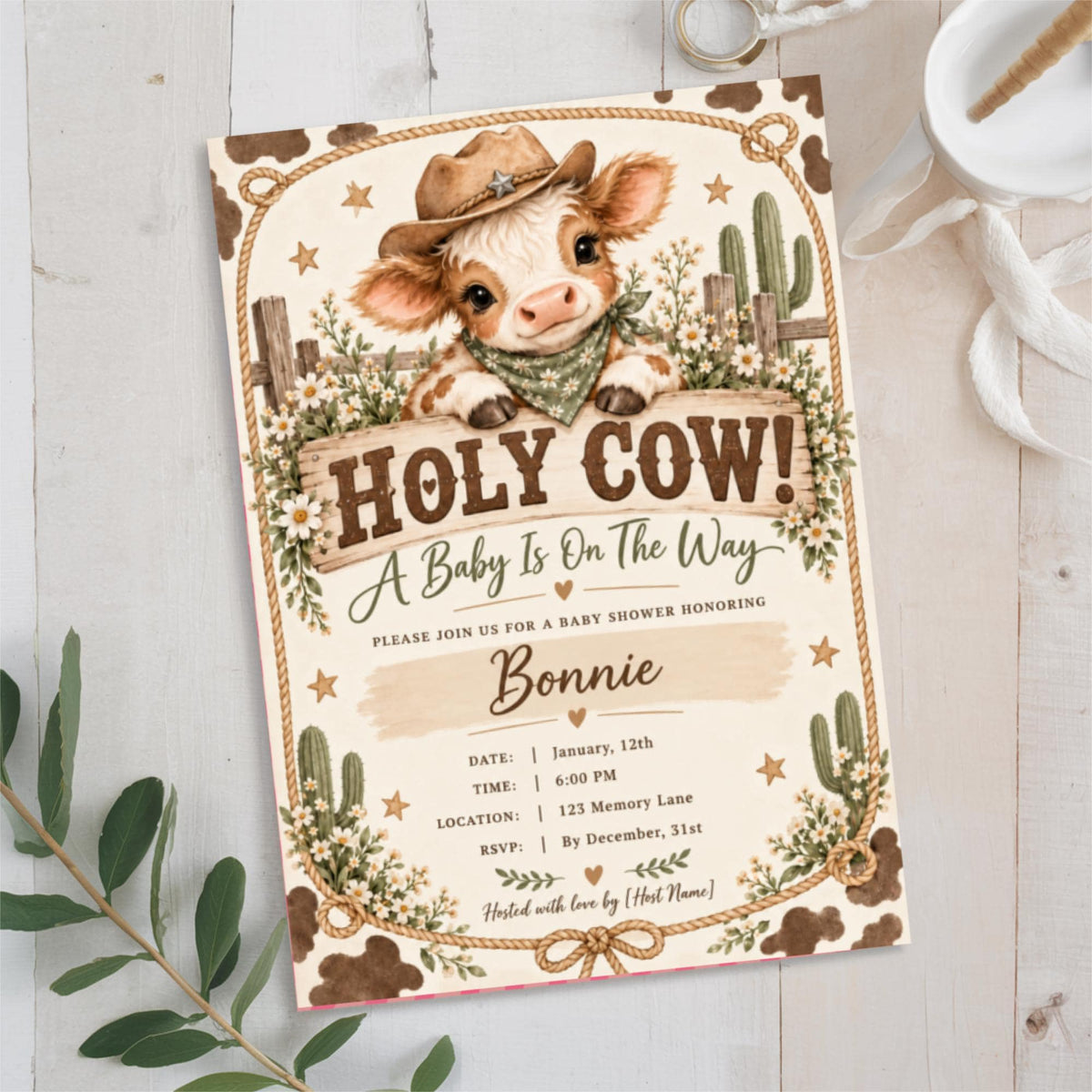 Holy Cow baby shower invitation displayed on a light neutral background with eucalyptus greenery. The rustic Western design features an adorable baby calf wearing a cowboy hat, white daisies, sage green accents, and decorative rope borders.