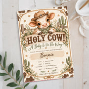 Holy Cow baby shower invitation displayed on a light neutral background with eucalyptus greenery. The rustic Western design features an adorable baby calf wearing a cowboy hat, white daisies, sage green accents, and decorative rope borders.