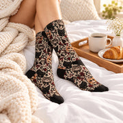 Woman lounging on white bedding wearing gothic cat socks featuring illustrated cats, burgundy roses, hearts, and black bows. A coffee mug, croissant, and chunky knit blanket complete the cozy scene.