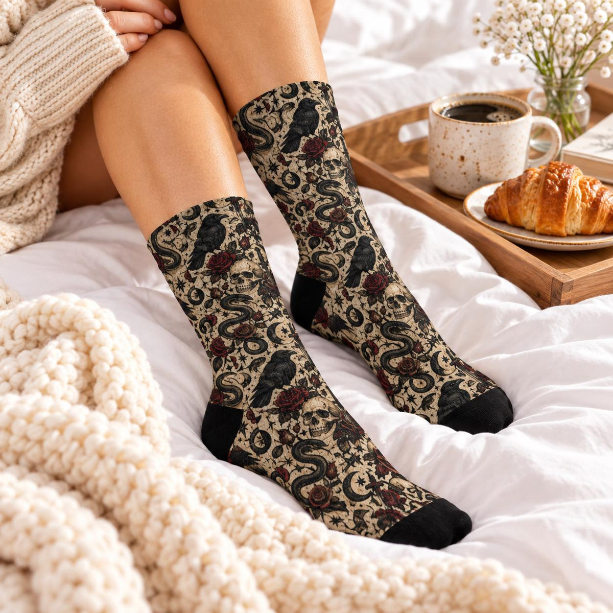 Gothic raven patterned crew socks displayed on a model relaxing in white bedding beside a coffee mug and croissant. Features dark bird illustrations, snakes, skulls, and red florals with black toes and heels.