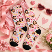 Pair of custom photo socks displayed with a pink gift box, satin ribbon, heart-shaped sunglasses, and flowers. Unique personalized gift featuring multiple faces and a dog against a playful pastel pink background.