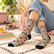 Playful retro dog crew socks worn with cuffed jeans in a cozy living room. Colorful design features dogs wearing sunglasses, hamburgers, hot dogs, smiley faces, lightning bolts, and vintage checkerboard accents.