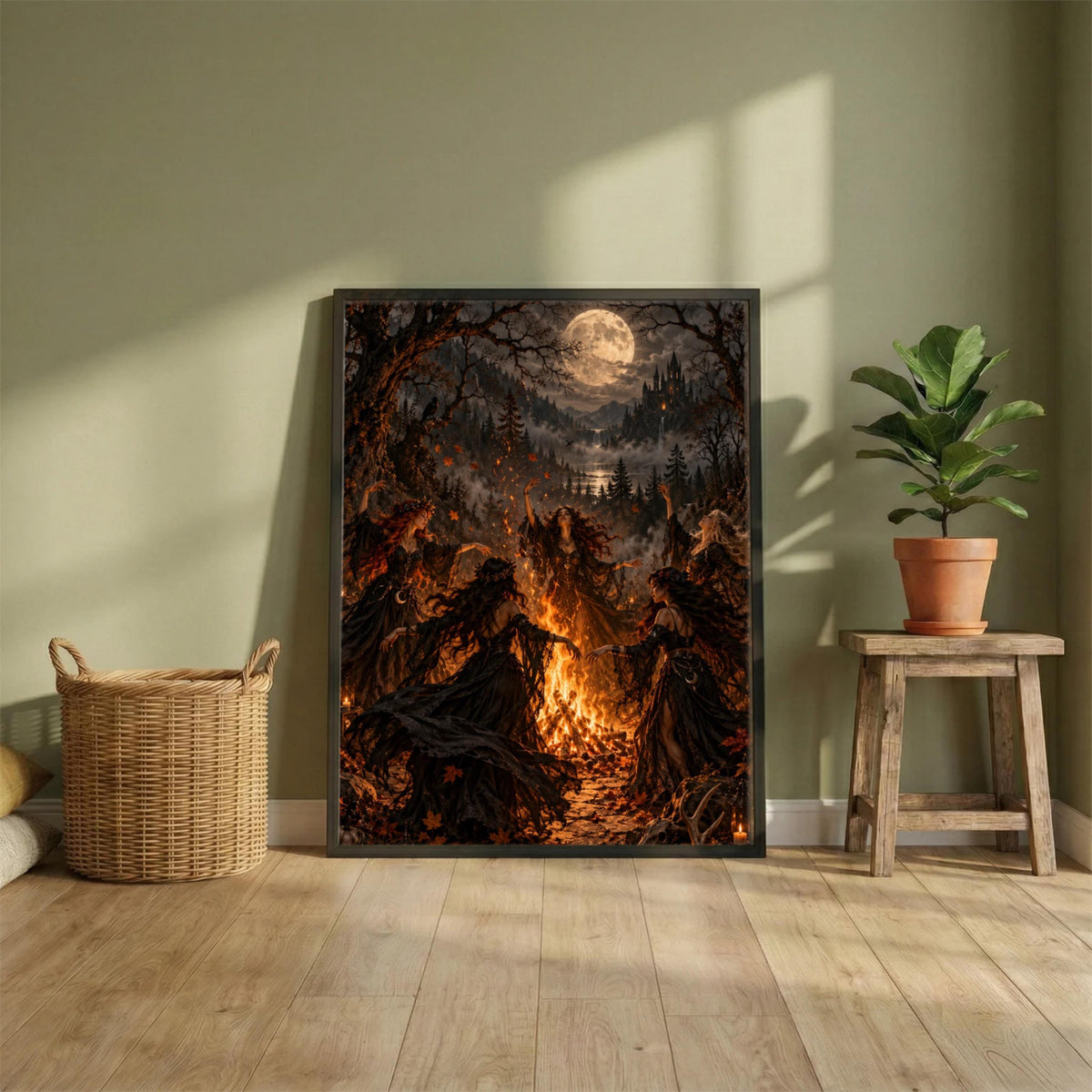 Framed witchy printable art resting on a wooden cabinet beside a large leafy plant in a sunlit neutral room. The print features a glowing bonfire, Gothic figures, and dark autumn woodland scenery.