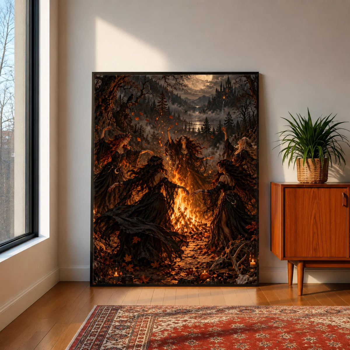 Dark cottagecore wall art resting on a wooden cabinet beside a leafy green houseplant. The dramatic artwork depicts five witches surrounding a fire with swirling autumn leaves and candlelit details.