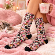 Retro cat crew socks worn by a woman relaxing on a fluffy pink blanket beside a sleeping kitten. Vintage-inspired pattern features playful cats, pink bows, leopard print accents, hearts, coffee cups, and black toes and heels.